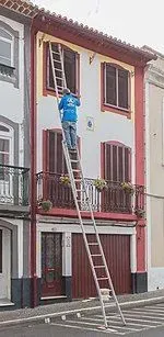 Persona subida a una escalera inspeccionando o reparando la fachada superior de un edificio blanco con molduras rojas.
